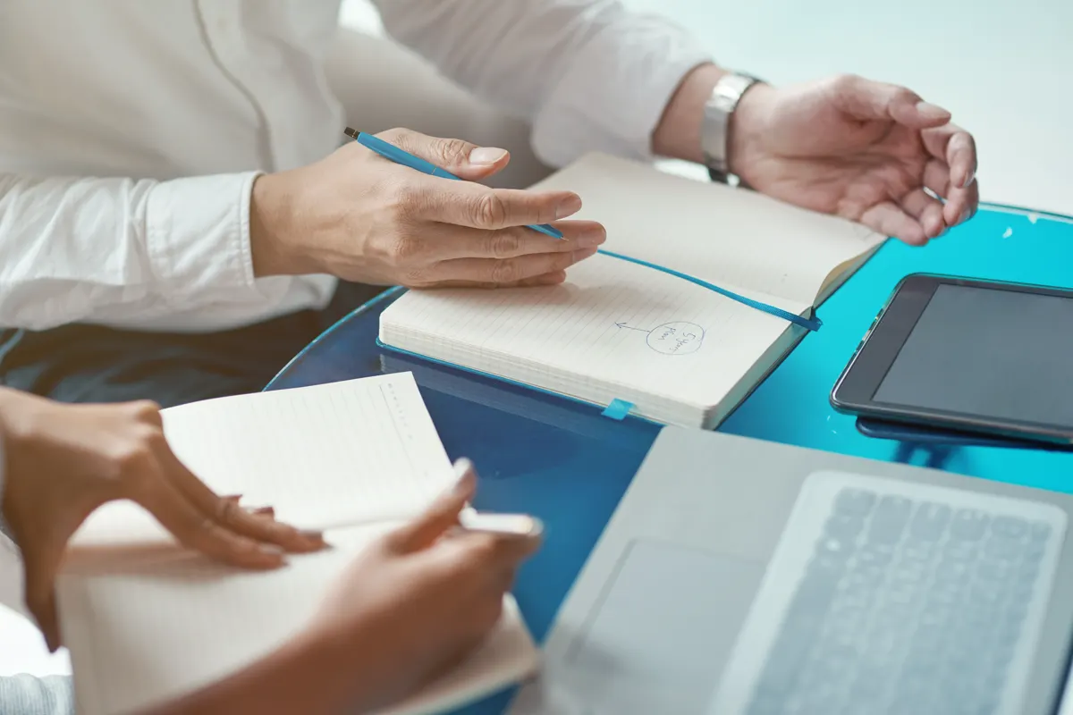 Hands, notebooks, and a tablet around a table in working discussion